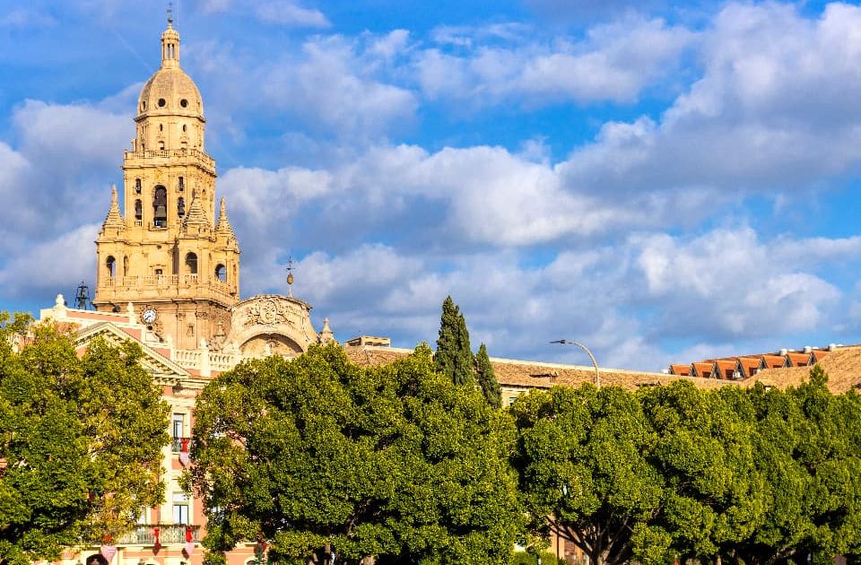Catedral de Murcia con su distintiva Torre Campanario de piedra dorada en la Ciudad de Murcia, rodeada de árboles y cielo azul.